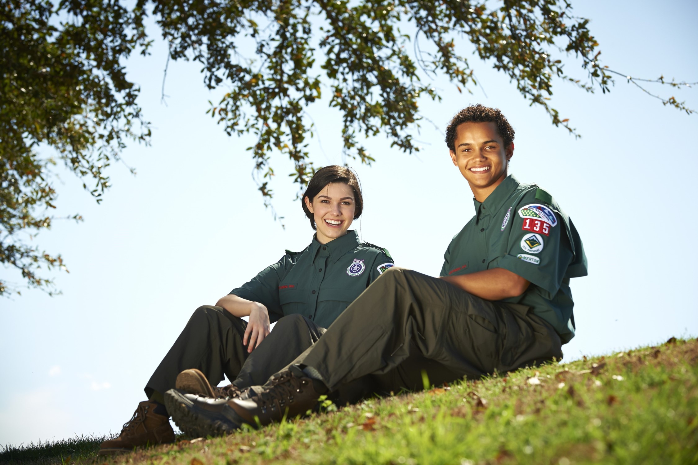Two older youth (around age 16), a man and a woman, wearing the Venturing uniform smile while sitting on a grassy hill