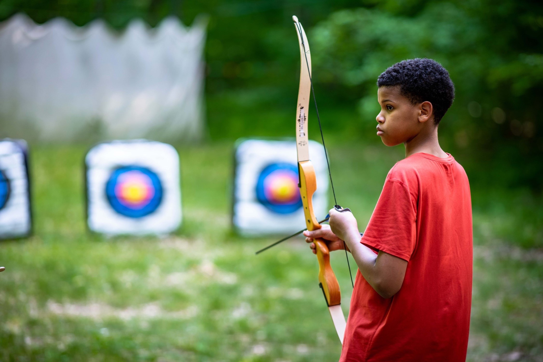 Young boy at an archery range holds a bow an arrow while looking to the side at the counselor, who is not visible in the photo