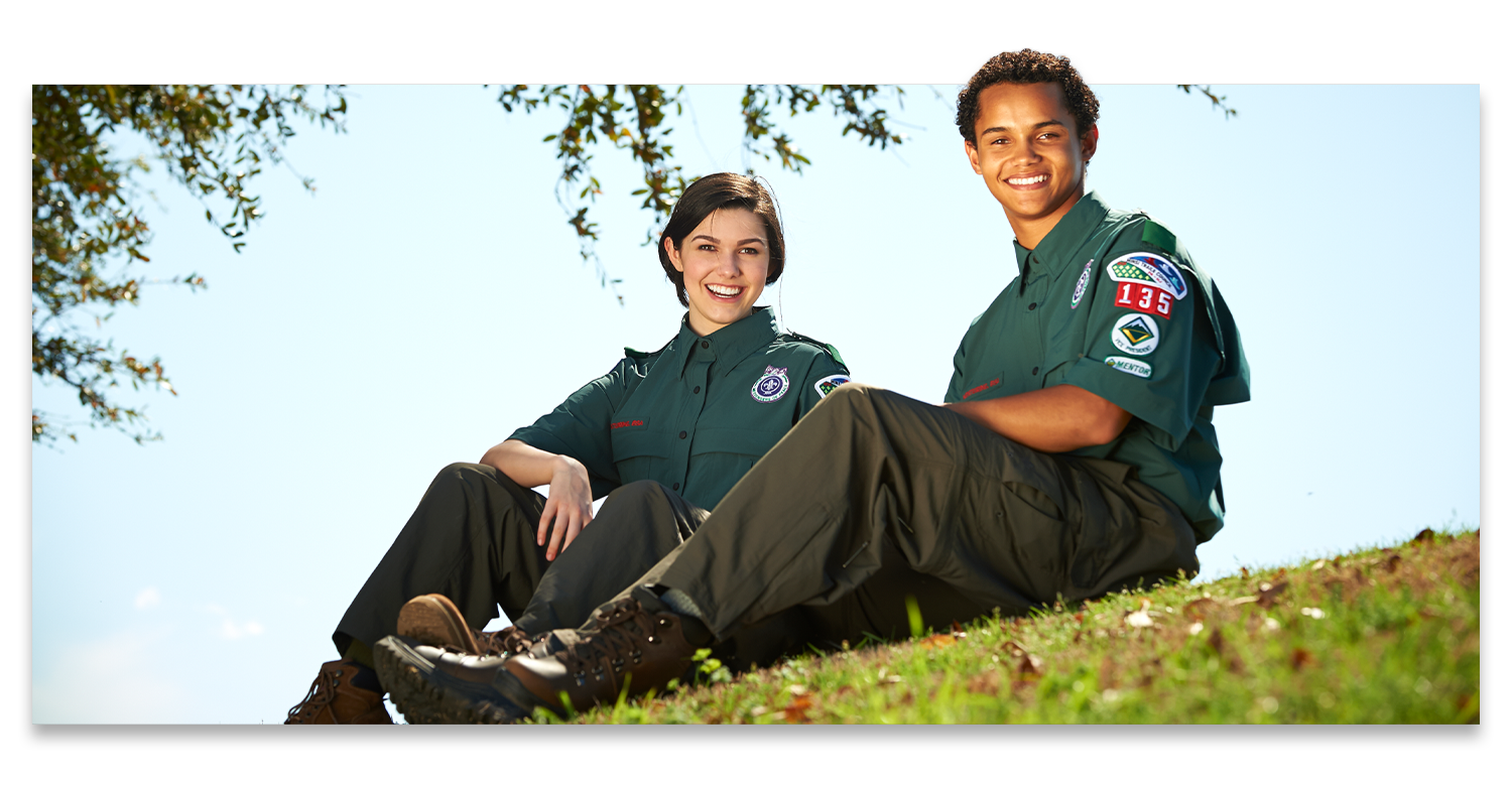 Two older teenagers smile while sitting outside wearing their venturing uniforms